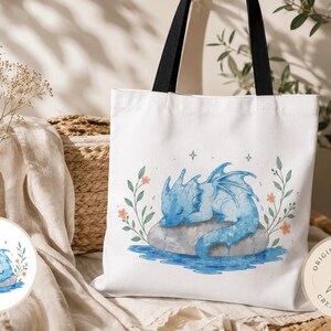 Könnte beinhalten: Eine weiße Tragetasche mit schwarzen Henkeln, die eine Aquarellillustration eines kleinen blauen Drachen zeigt, der auf einem grauen Stein im blauen Wasser schläft, umgeben von grünen Blattstielen mit kleinen orangefarbenen Blüten und Sternen.