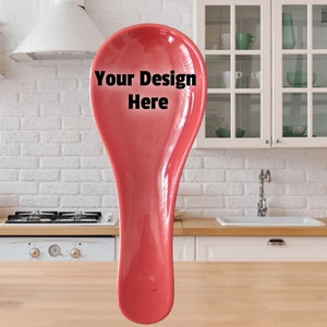 May include: A vibrant red ceramic spoon rest with the text "Your Design Here" in black. The spoon rest has a curved, elongated design. The background shows a kitchen setting with white cabinets and a brick wall.