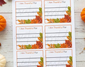 May include: A white paper with six sections, each labeled "I Am Thankful For." The sections have horizontal lines for writing and are decorated with orange, green, and red autumn leaves. The image is surrounded by fall foliage and acorns.