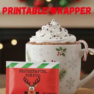 Peut inclure: Une tasse de chocolat chaud garnie de crème fouettée et de copeaux de chocolat, avec un sucre d'orge. À côté, un sachet rouge et vert de mélange de chocolat chaud "Reindeer Fuel for Fun". Le texte "Hot Chocolate Printable Wrapper" est également présent.