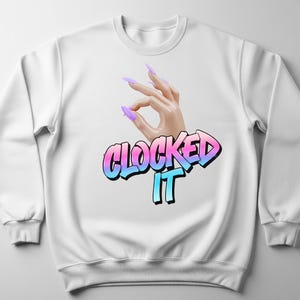 Op de afbeelding: Witte sweatshirt met een hand die het 'oké'-gebaar maakt, versierd met lange, paarse nagels. De woorden 'CLOCKED IT' zijn geschreven in een blauwe en roze gradiënt, omlijnd in zwart.