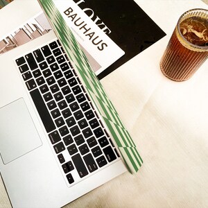 Puede incluir: Un ordenador port&aacute;til plateado con un teclado negro y una funda a rayas verde y blanca. Un vaso de t&eacute; helado con una pajita se encuentra sobre una mesa blanca.