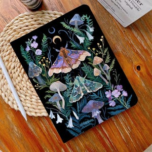 May include: A black notebook cover featuring a whimsical illustration of moths, mushrooms and flowers on a dark background. The illustration includes a large moth with purple and brown wings, a smaller green moth, and various mushrooms in shades of white, brown and purple.