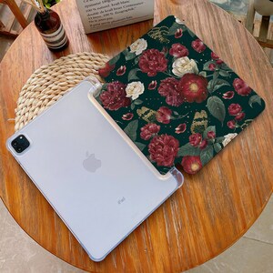 May include: A white iPad case with a floral design. The case has a dark green background with red, white, and gold flowers. The iPad is visible in the background.