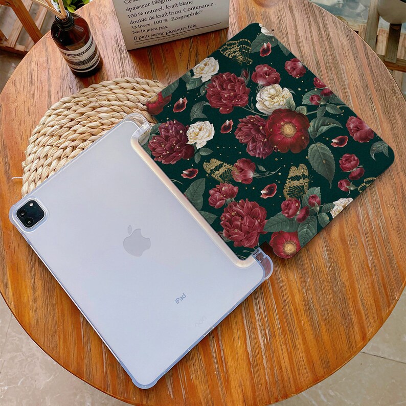 May include: A white iPad case with a floral design. The case has a dark green background with red, white, and gold flowers. The iPad is visible in the background.