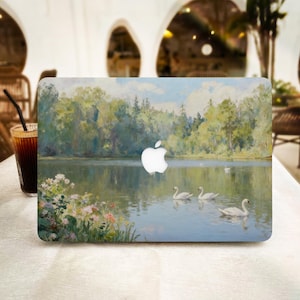 May include: A laptop with a scenic landscape design featuring a lake, trees, and swans. The artwork has a vintage aesthetic with soft colors and a serene atmosphere. The Apple logo is visible.