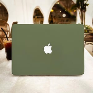 Op de afbeelding: Olijfgroene laptop met een wit Apple-logo op een witte tafel. Links van de laptop staat een glas ijskoffie met een rietje. De achtergrond is een wazig beeld van een café.