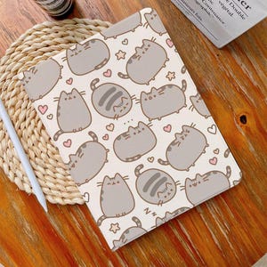 Puede incluir: Una funda para iPad de color blanco y crema con un patrón repetido de gatos de dibujos animados grises con corazones y estrellas. La funda tiene un acabado suave y texturizado.