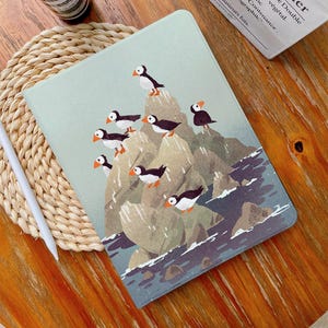 May include: A light green tablet case with a charming illustration of puffins on rocky cliffs. The artwork shows the birds with orange beaks and black and white feathers, set against a backdrop of sea and sky. The case is designed to protect a tablet.