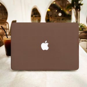 Puede incluir: Un portátil marrón con el logotipo de Apple en la tapa se muestra sobre una mesa blanca. Un vaso de café helado con una pajita está a la izquierda del portátil. El fondo está borroso, mostrando un ambiente de cafetería.