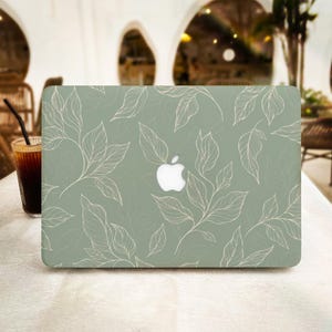 Puede incluir: Una funda para portátil de color verde salvia con un delicado patrón de líneas blancas de hojas dispersas por toda la superficie. El diseño incluye un recorte central que revela el logotipo de Apple blanco iluminado en la tapa.