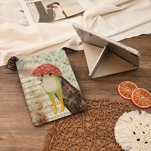 Puede incluir: Una funda para tableta con una ilustración de una rana con un sombrero de seta y botas amarillas, sobre un fondo de partituras y motivos florales. Una tableta transparente está apoyada en un soporte plateado. También se ven rodajas de naranja decorativas y un posavasos de macramé.