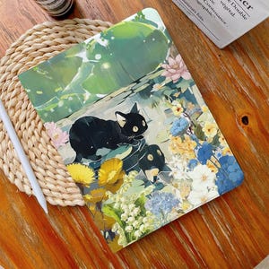 May include: A black cat illustration on a tablet case. The case features a colorful floral design with yellow, blue, and white flowers. The background has a green and light blue watercolor effect. A white stylus rests on a woven mat.