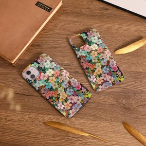 Op de afbeelding: Twee telefoonhoesjes met een bloemenpatroon. De hoesjes hebben een kleurrijk scala aan kleine bloemen in roze, blauwe, gele en oranje tinten. De hoesjes liggen op een houten oppervlak.