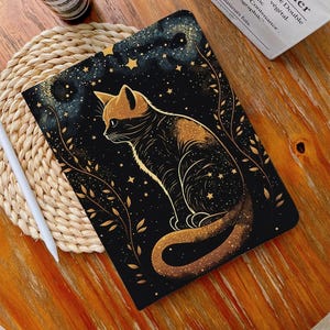 Puede incluir: Funda negra para tableta con una ilustración estilizada de un gato con detalles dorados. El gato está sobre un cielo estrellado con estrellas y follaje dorado. Un lápiz óptico blanco descansa sobre una esterilla tejida.