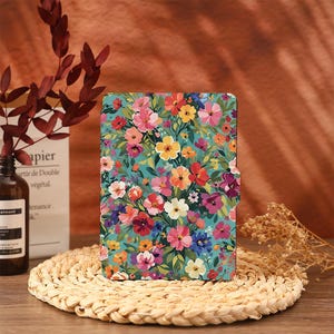 Puede incluir: Una funda para tableta con estampado floral, fondo turquesa y una vibrante variedad de flores rosas, rojas, amarillas y moradas. La funda se cierra con un cierre magnético. La funda está sobre un salvamanteles tejido.