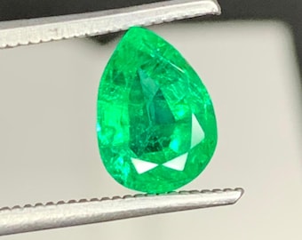 Puede incluir: Una piedra preciosa de esmeralda facetada en forma de pera con un color verde vibrante y saturado. La piedra está sujeta por unas pinzas de metal plateado, mostrando su claridad interna, bordes afilados y superficies pulidas.