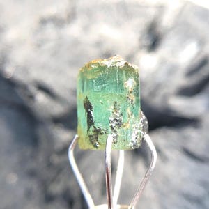 May include: A green emerald gemstone with a rough, natural finish. The emerald is held in a metal clamp.