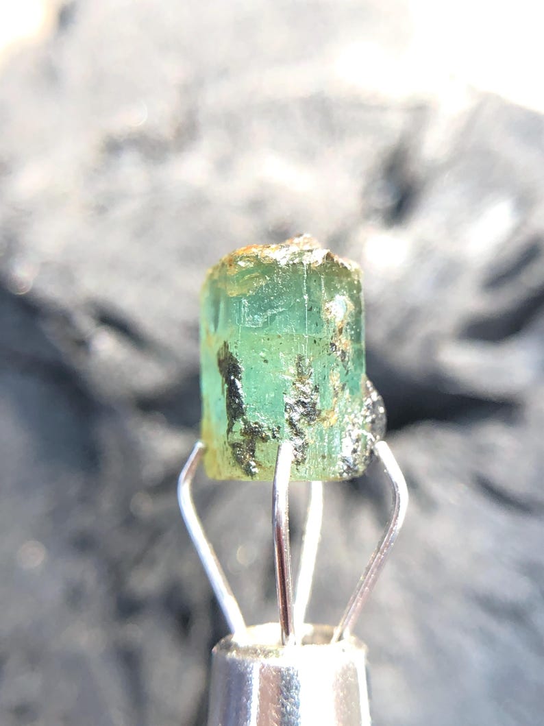 May include: A green emerald gemstone with a rough, natural finish. The emerald is held in a metal clamp.