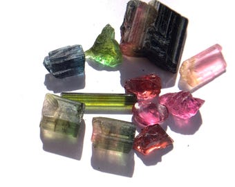 May include: A collection of raw tourmaline crystals in various colors, including green, pink, blue, and black. The gemstones are displayed on a white surface, showcasing their natural shapes and textures. The crystals vary in size and shape, with some appearing translucent.