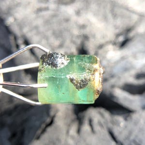 May include: A green emerald gemstone with a rough, natural finish. The emerald is held in a metal clamp.