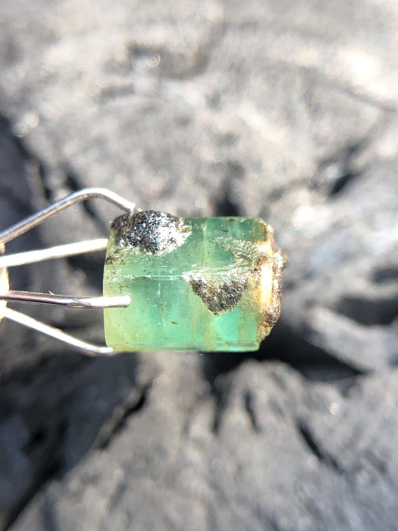 May include: A green emerald gemstone with a rough, natural finish. The emerald is held in a metal clamp.