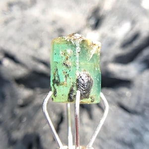 May include: A green emerald crystal with black inclusions, held in a metal clamp.