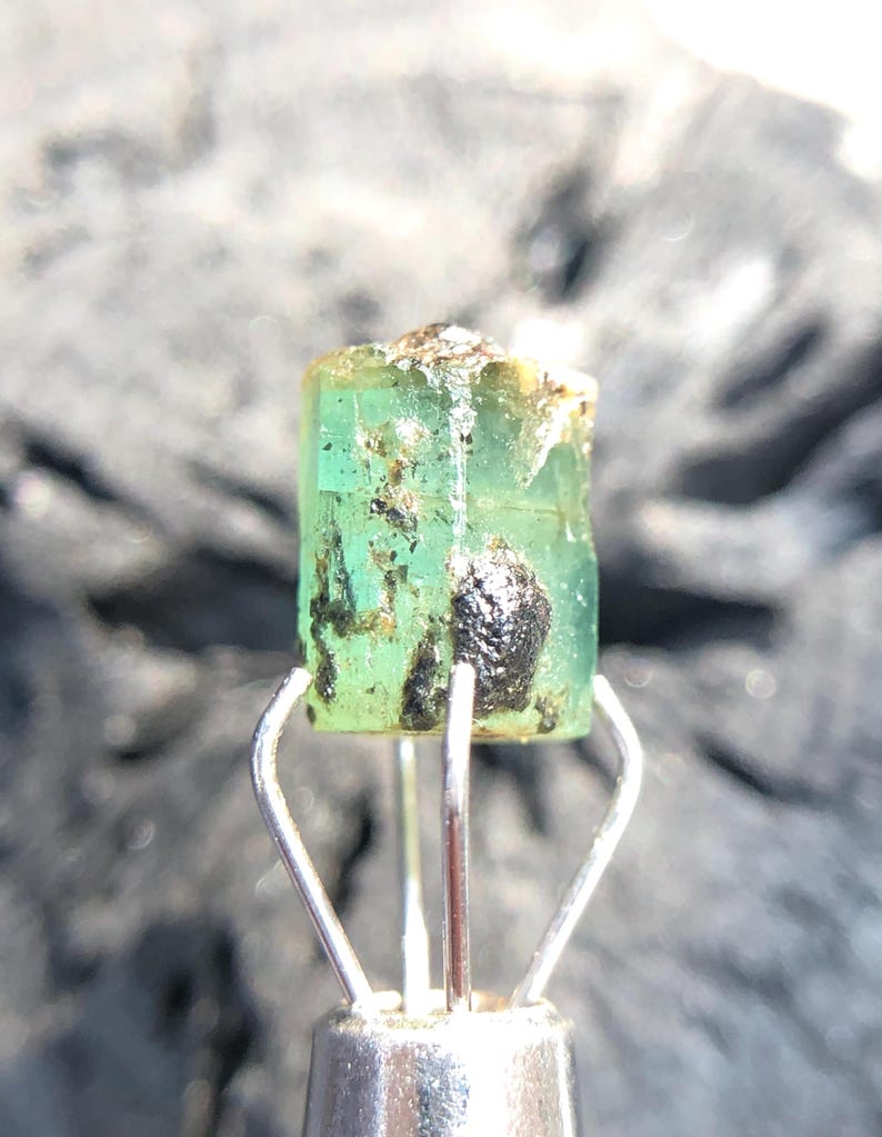 May include: A green emerald crystal with black inclusions, held in a metal clamp.