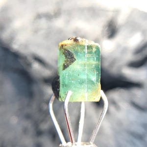 May include: A green emerald gemstone with black inclusions, held in a metal clamp.