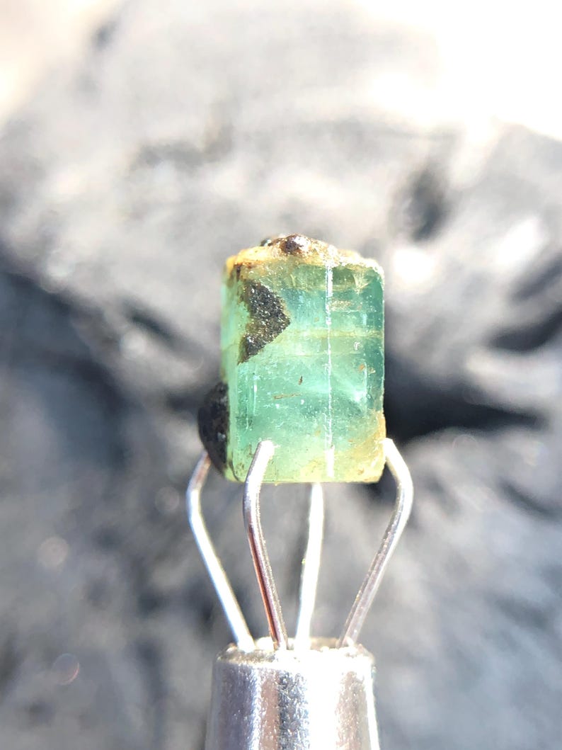 May include: A green emerald gemstone with black inclusions, held in a metal clamp.