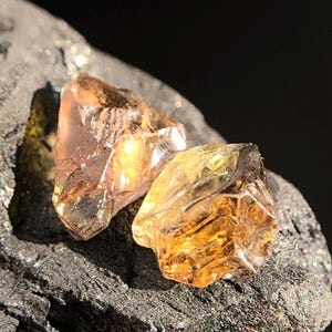 Könnte beinhalten: Zwei grob geschnittene, bernsteinfarbene Edelsteine mit kristalliner Struktur. Die Edelsteine liegen auf einer dunklen, strukturierten Oberfläche mit schwarzem Hintergrund. Die Edelsteine haben einen warmen, goldenen Farbton und reflektieren Licht.