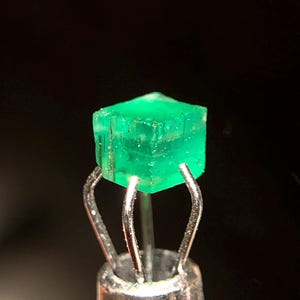 May include: A vivid, emerald-green gemstone, cut into a cube shape, is held by a silver metal tool. The gemstone's facets reflect light, showcasing its clarity. The background is a dark, solid colour, emphasising the gem's brilliance.
