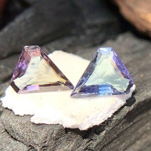 May include: Two triangular faceted gemstones, one with a purple and green hue and the other with a blue and green hue, are displayed on a white surface.