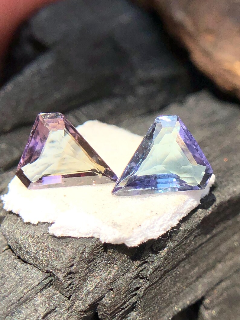 May include: Two triangular faceted gemstones, one with a purple and green hue and the other with a blue and green hue, are displayed on a white surface.
