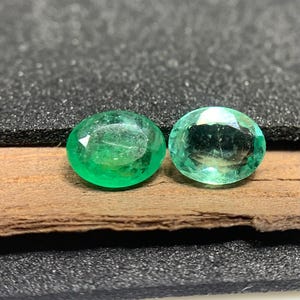 Può includere: Due pietre preziose di smeraldo taglio ovale. Le gemme sono di un colore verde vibrante e sembrano sfaccettate. Sono esposte su un pezzo di legno, con uno sfondo scuro.