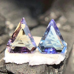 May include: Two triangular faceted gemstones, one purple and yellow, the other blue and purple, on a white surface.