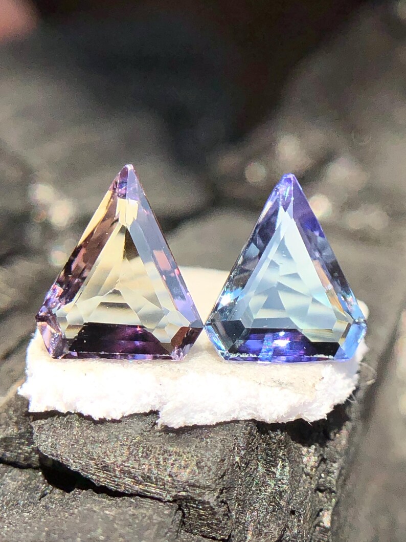 May include: Two triangular faceted gemstones, one purple and yellow, the other blue and purple, on a white surface.