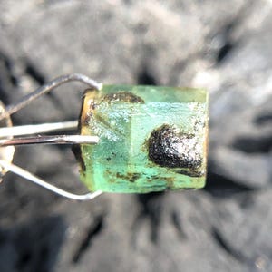 May include: A green emerald gemstone with black inclusions, held in a metal clamp.