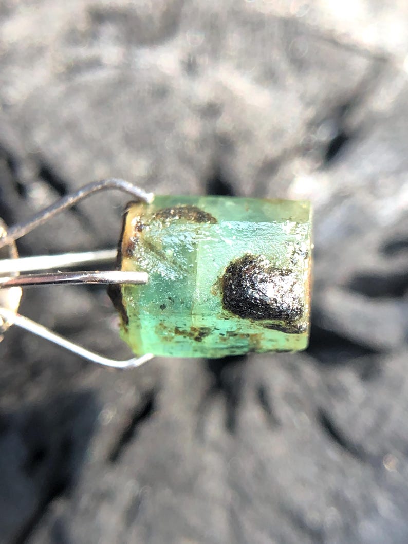 May include: A green emerald gemstone with black inclusions, held in a metal clamp.