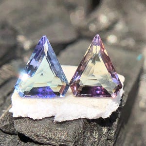 May include: Two triangular gemstones, one with a blue-green hue and the other with a yellow-green hue, both with a purple tint. The gemstones are set on a white surface.