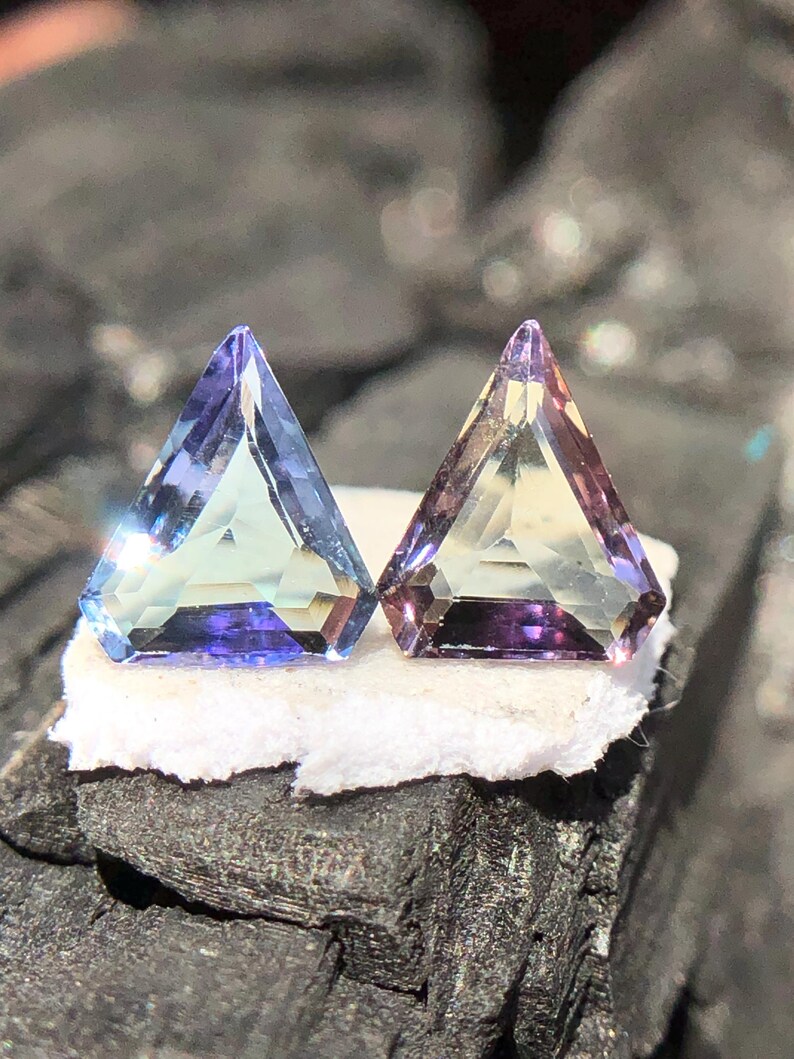 May include: Two triangular gemstones, one with a blue-green hue and the other with a yellow-green hue, both with a purple tint. The gemstones are set on a white surface.