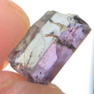 May include: A rectangular, faceted gemstone with a translucent appearance, displaying a gradient of pale lavender and purple hues. The stone features internal inclusions and natural fractures, with a clear, glassy texture and sharp, defined edges.