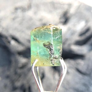 May include: A green emerald gemstone with a rectangular shape and a slight golden hue. The gemstone is held in a pair of tweezers.