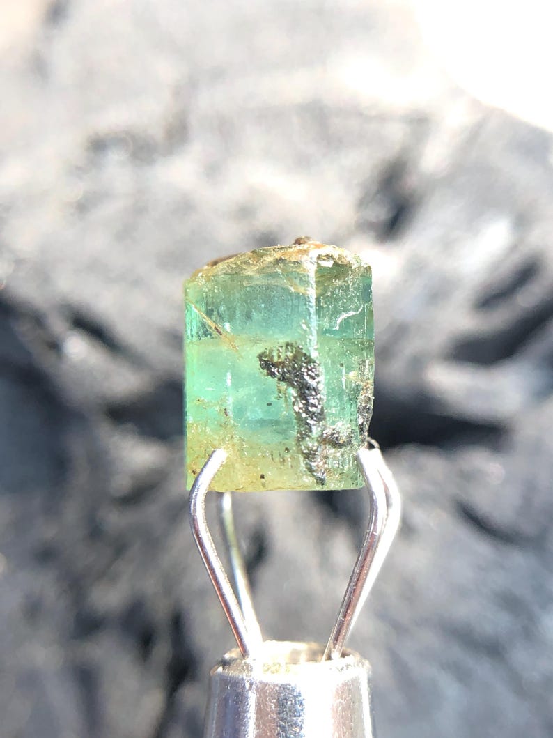 May include: A green emerald gemstone with a rectangular shape and a slight golden hue. The gemstone is held in a pair of tweezers.