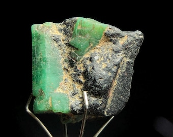 May include: A raw emerald gemstone with a vibrant green hue, partially encased in dark, rough matrix. The emerald is held by a silver metal stand against a black background, showcasing its natural texture and color.