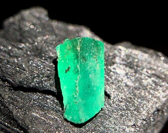 May include: A vibrant, raw emerald gemstone with a rich green hue. The rough-cut crystal displays natural facets and textures, set against a dark, textured background. This gemstone is a striking example of natural mineral beauty.