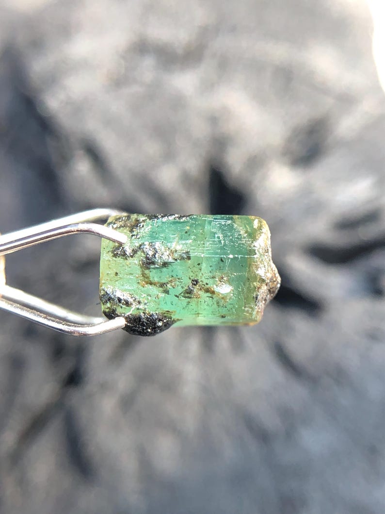 May include: A rough, green emerald gemstone with black inclusions. The emerald is held in a pair of tweezers.