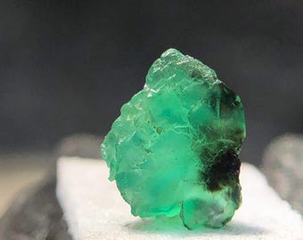 May include: A rough-cut, vibrant green emerald gemstone. The raw mineral displays a crystalline structure with a dark inclusion. The emerald is set on a white, textured surface against a dark background.