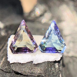 May include: Two triangular gemstones, one purple and one blue, with a faceted cut. The gemstones are set on a white surface.