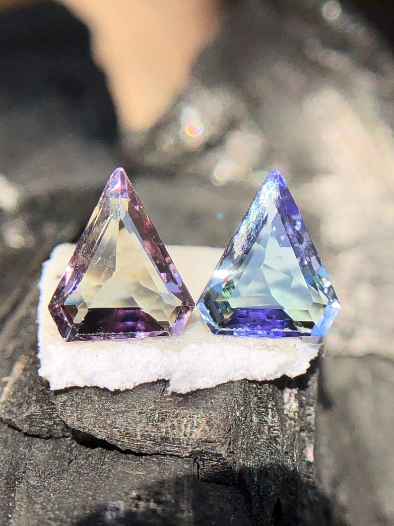 May include: Two triangular gemstones, one purple and one blue, with a faceted cut. The gemstones are set on a white surface.